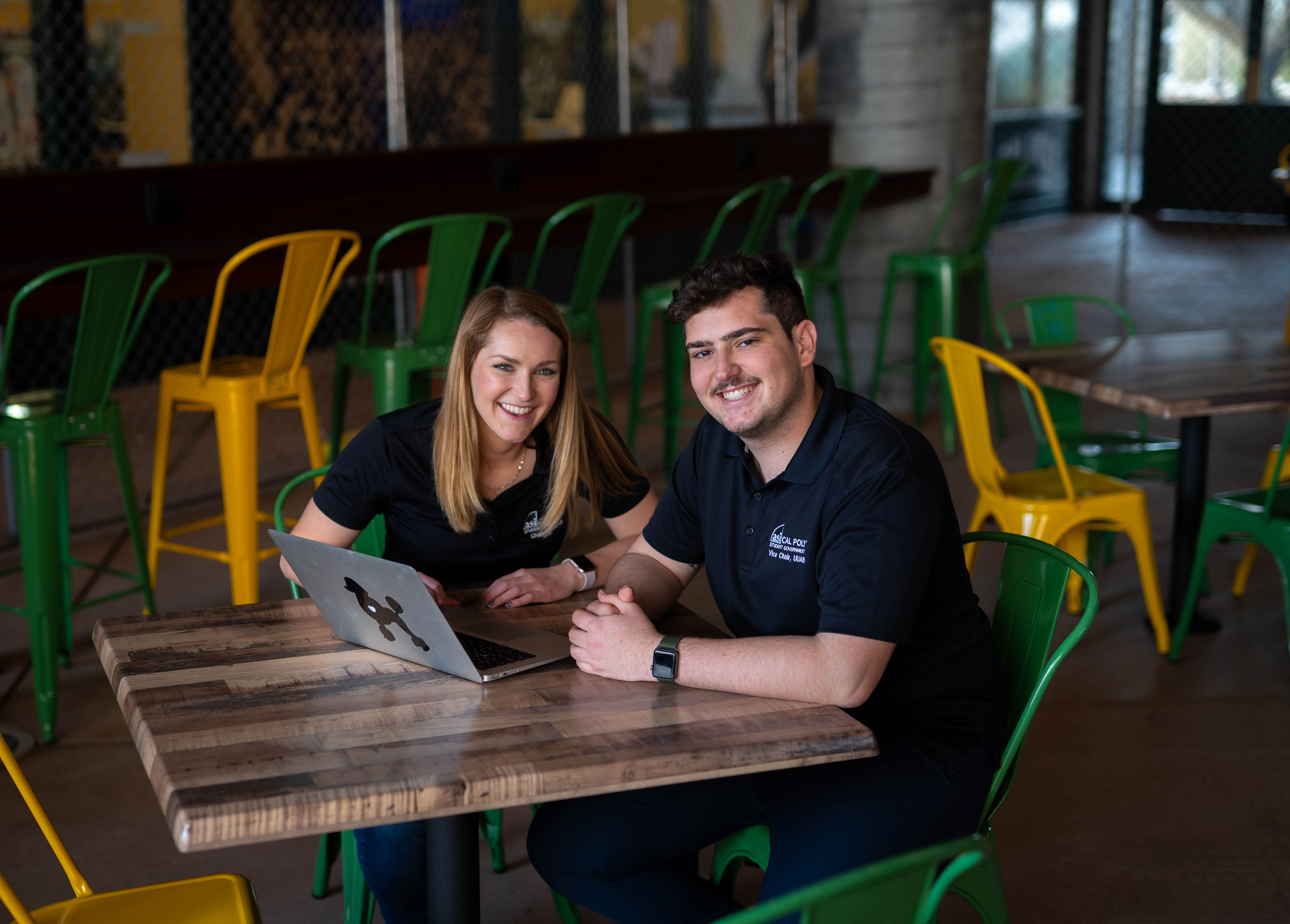 Elizabeth Roseman and Will Sambar sit smiling in green chairs at a wood table with a laptop on it 